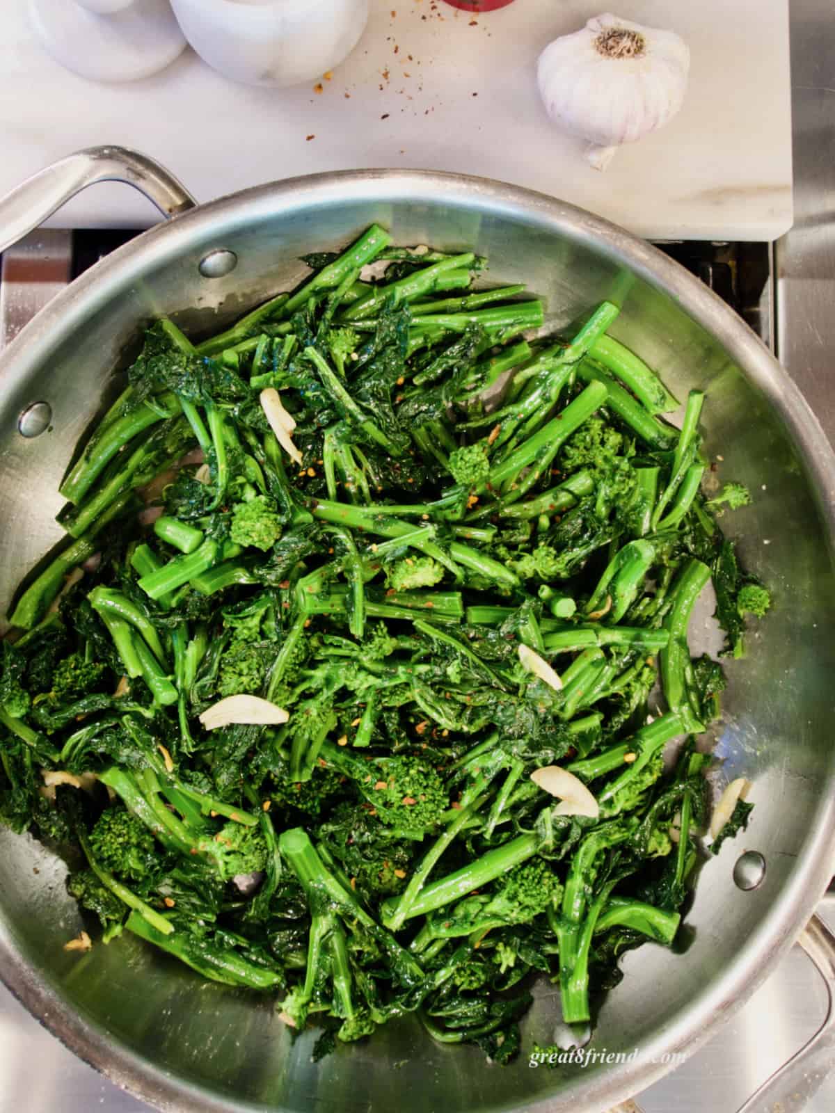 Overhead shot of cooked broccoli rabe in a stainless steel pan with slices of garlic and a head of garlic next to the pan.