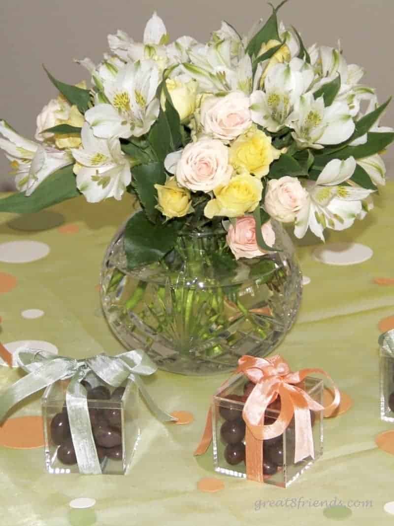 A round glass vase filled with fresh flowers in light pastel colors sitting on a light green tablecloth with small clear boxes of chocolates.