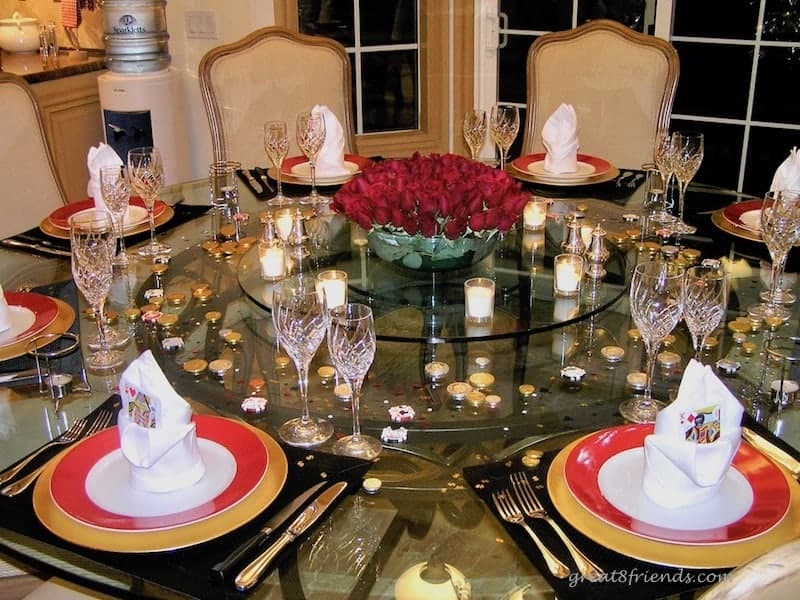 A round table set for a Las Vegas Dinner Party with a centerpiece of red roses surrounded by votive candles. foil wrapped chocolates are on the table and the plates are red rimmed sitting on gold chargers.