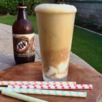 One tall root beer float with a bottle of A&W in the background and several colorful straws laying in front.