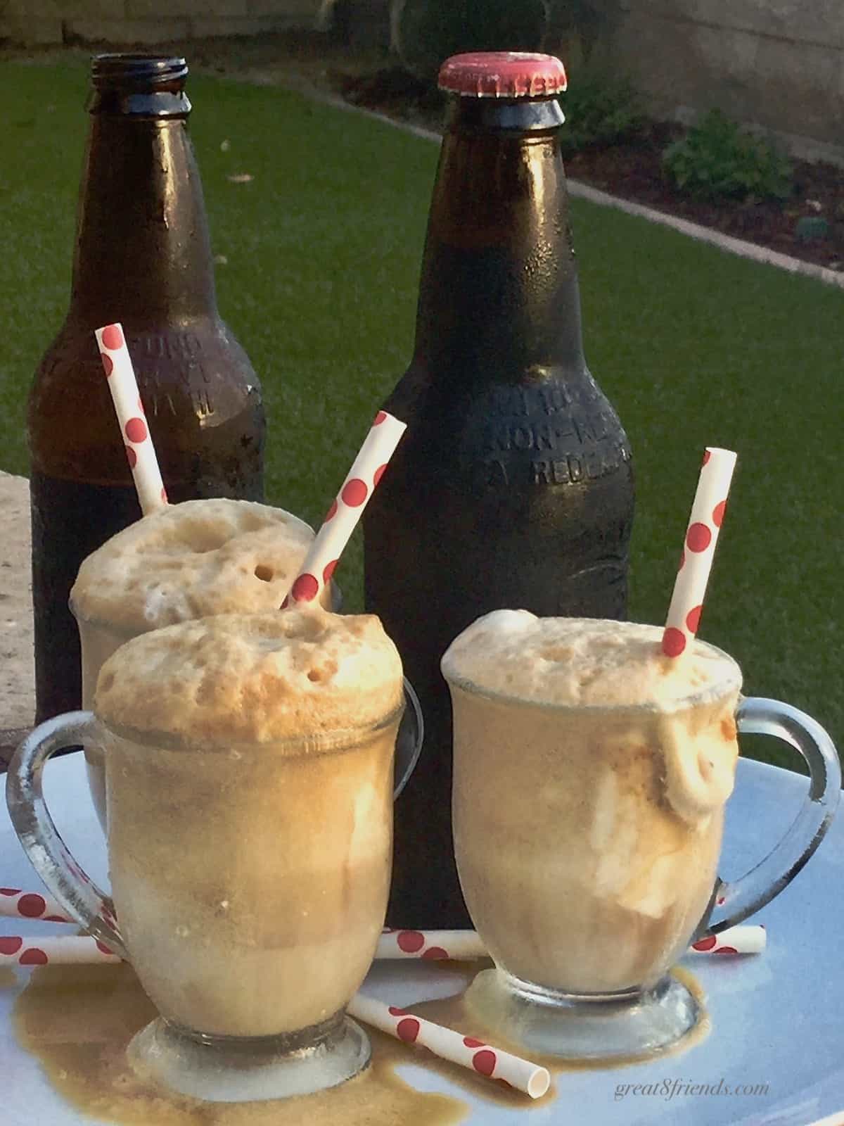 Three glass mugs filled with root beer floats with straws and 2 brown bottles in the background.