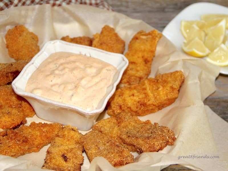 Several small pieces of catfish served on brown paper with a sauce in a small bowl in the middle with several lemon wedges on the side.