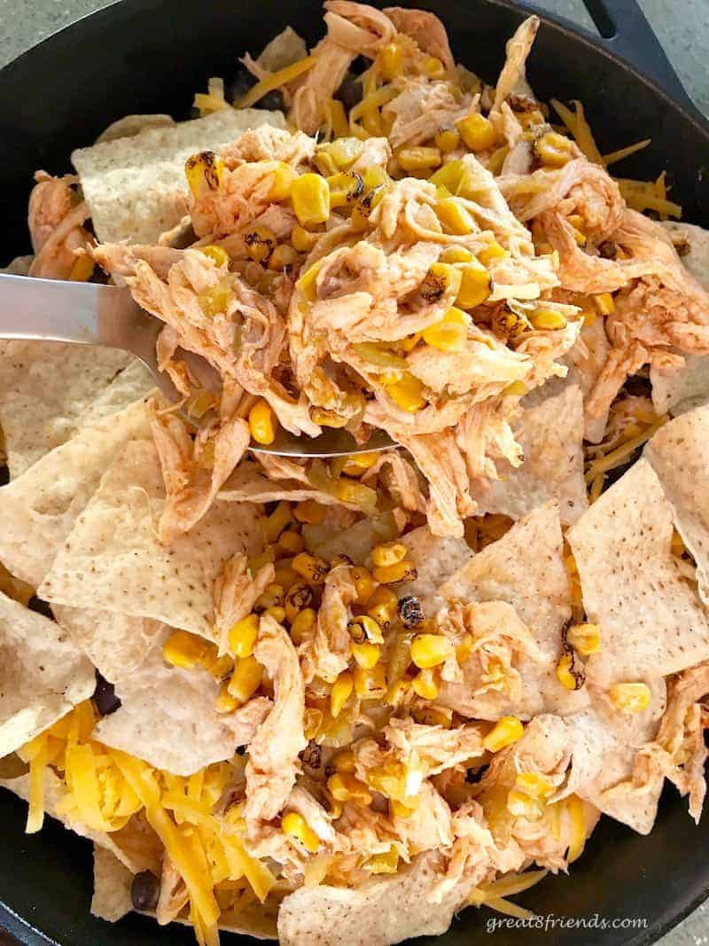 A large spoon adding chicken mixture to skillet of chips for nachos.