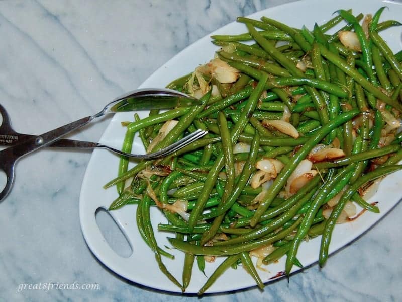 Overhead shot of green beans and onions on a white platter. with tongs to serve.