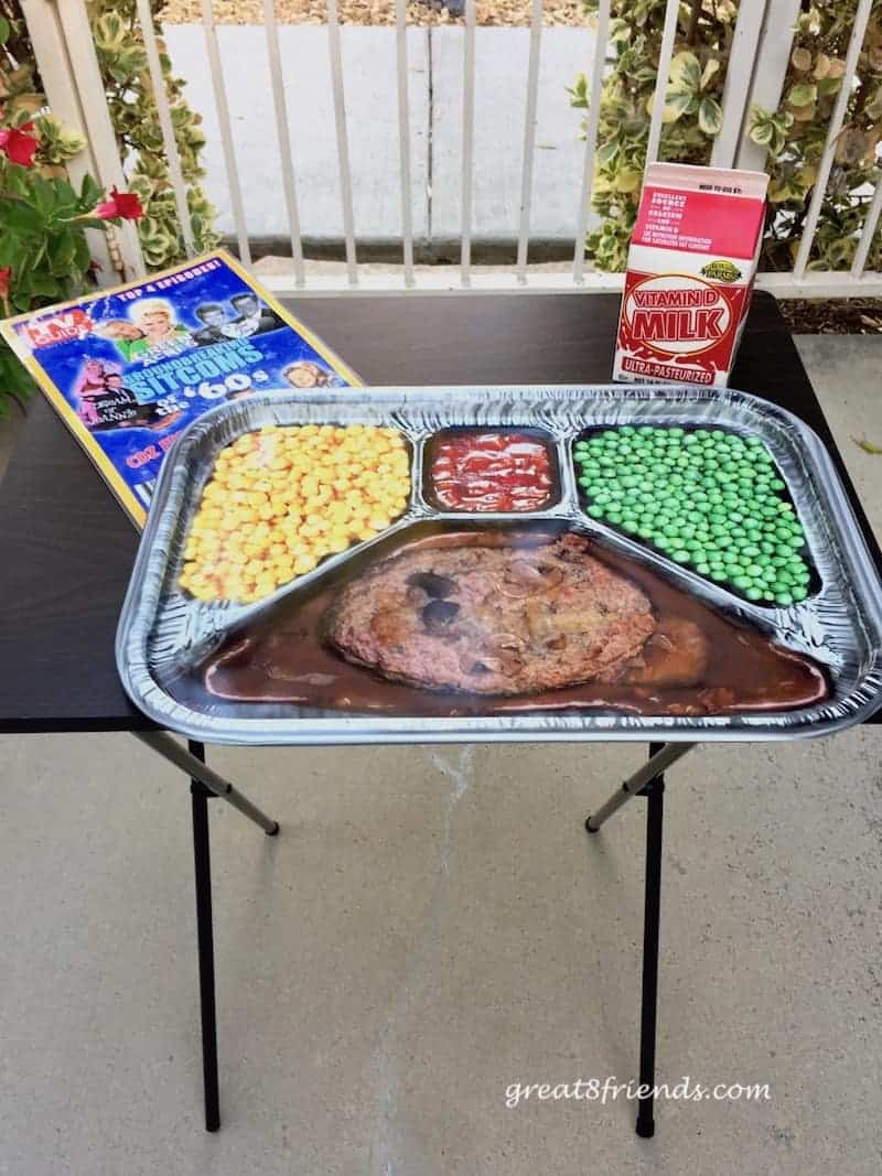 A tv tray with fake tv dinner, mocked up TV guide and carton of milk.