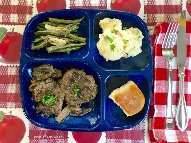 Blue divided plate with salisbury steak, green beans, mashed potatoes and a dinner roll.