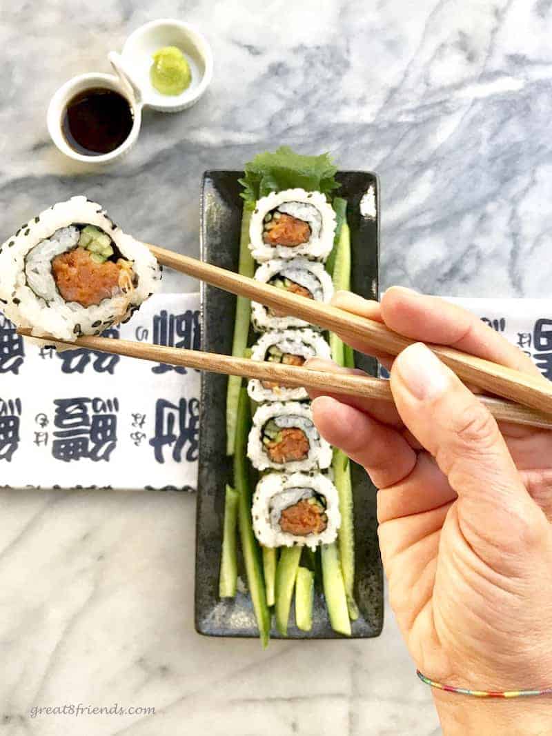 A piece of sushi being held between chopsticks with the tray of sushi behind.