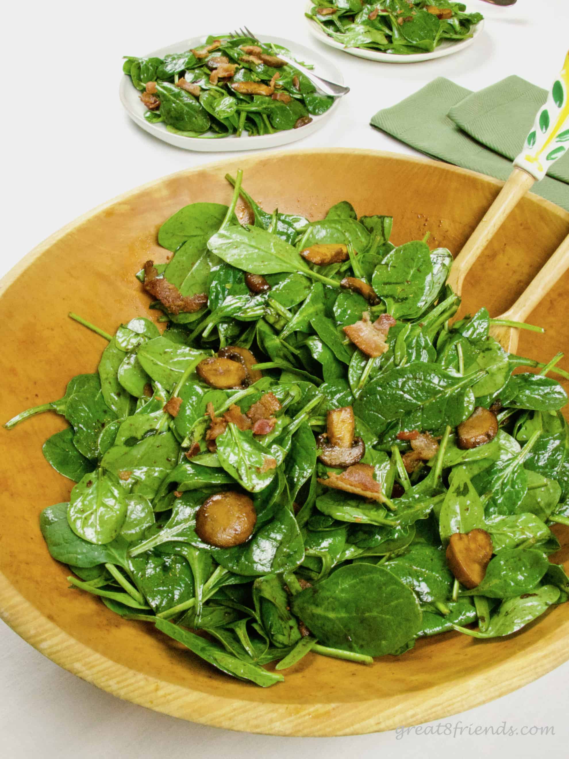 Spinach salad with mushrooms and bacon in a wooden bowl with plates of salad in the background.