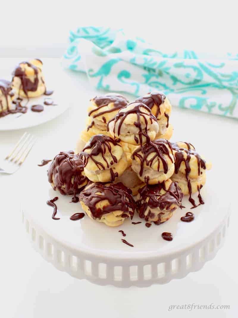 Several homemade Profiteroles with ice cream and chocolate sauce stacked in the center of a white cake plate.