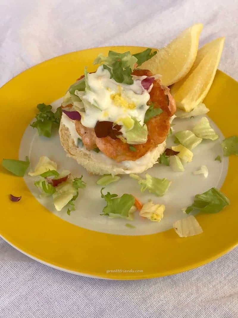 Salmon Slider with Tangy Lemony Yogurt Sauce served on a bottom bun only on a yellow plate with two lemon wedges.