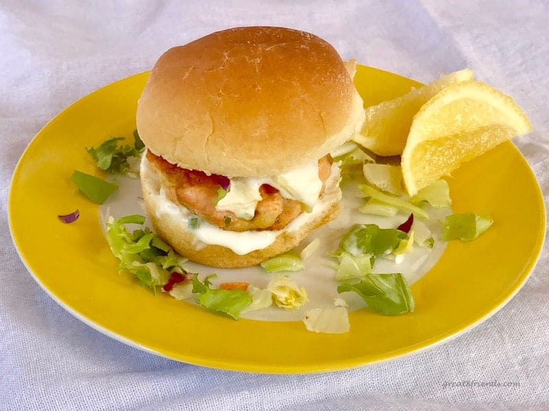 Salmon Slider with Tangy Lemony Yogurt Sauce served on a bun on a yellow plate with two lemon wedges on the side.