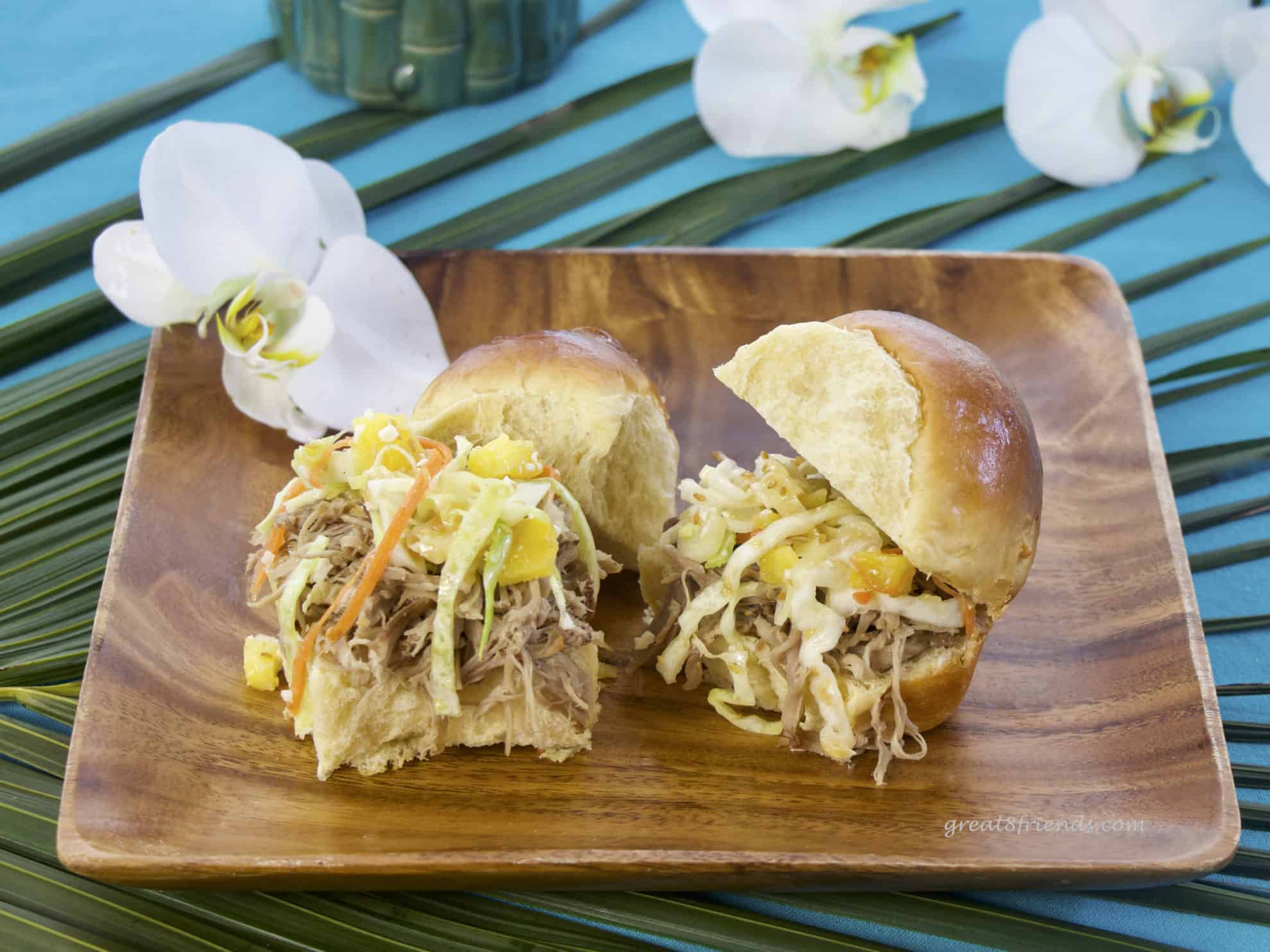 Two pulled pork sandwiches on Hawaiian sweet rolls sitting on a square wooden plate garnished with an orchid.