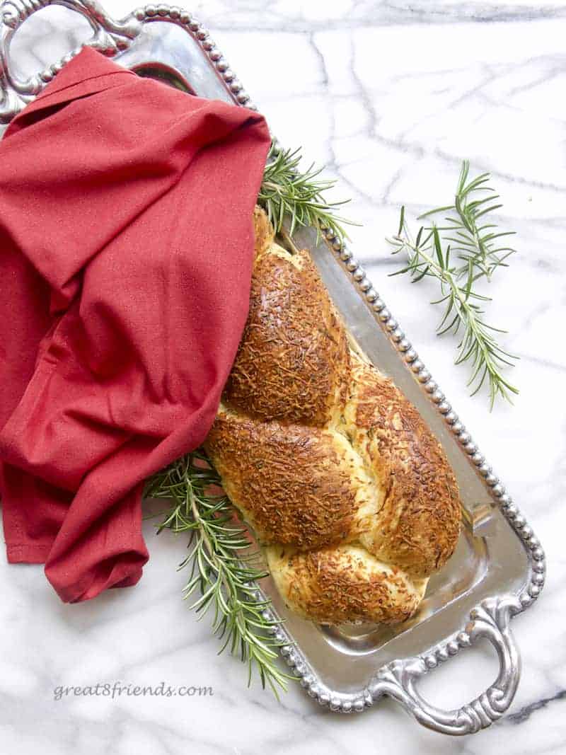 A loaf of braided cheese bread on a silver tray with branches of rosemary laying around and a red napkin.