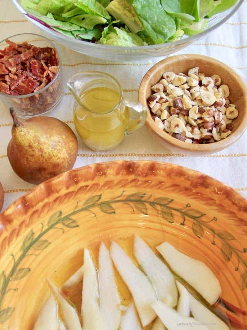 Overhead view of a large salad bowl with sliced pears in the bottom marinating in salad dressing with a pear, pitcher of dressing and chopped hazelnuts on the side.