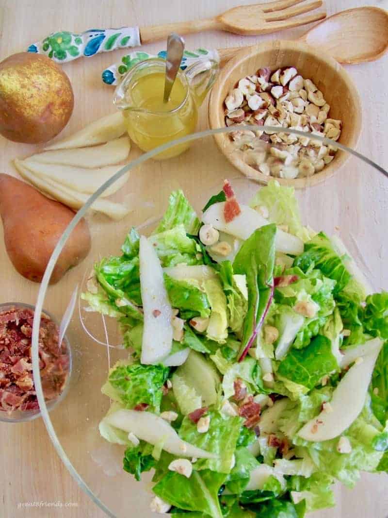 Overhead photo of a green salad in a glass bowl with hazelnuts, bacon bits and sliced and whole pears on the side.