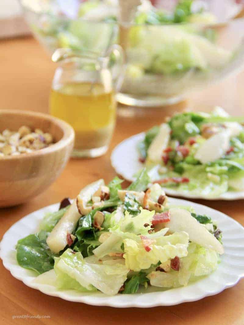 This unclose photo of a white plate of salad with green lettuce with pears and bacon bits and hazelnuts sprinkled on top with a pitcher of dressing in the background.