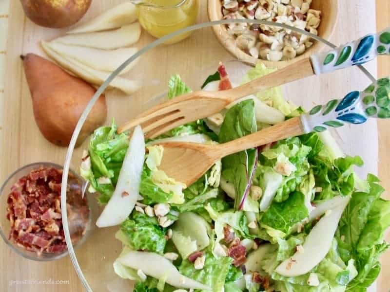 Salad with wood and ceramic salad tossers on the top and on the side is chopped bacon, pears, and hazelnuts.