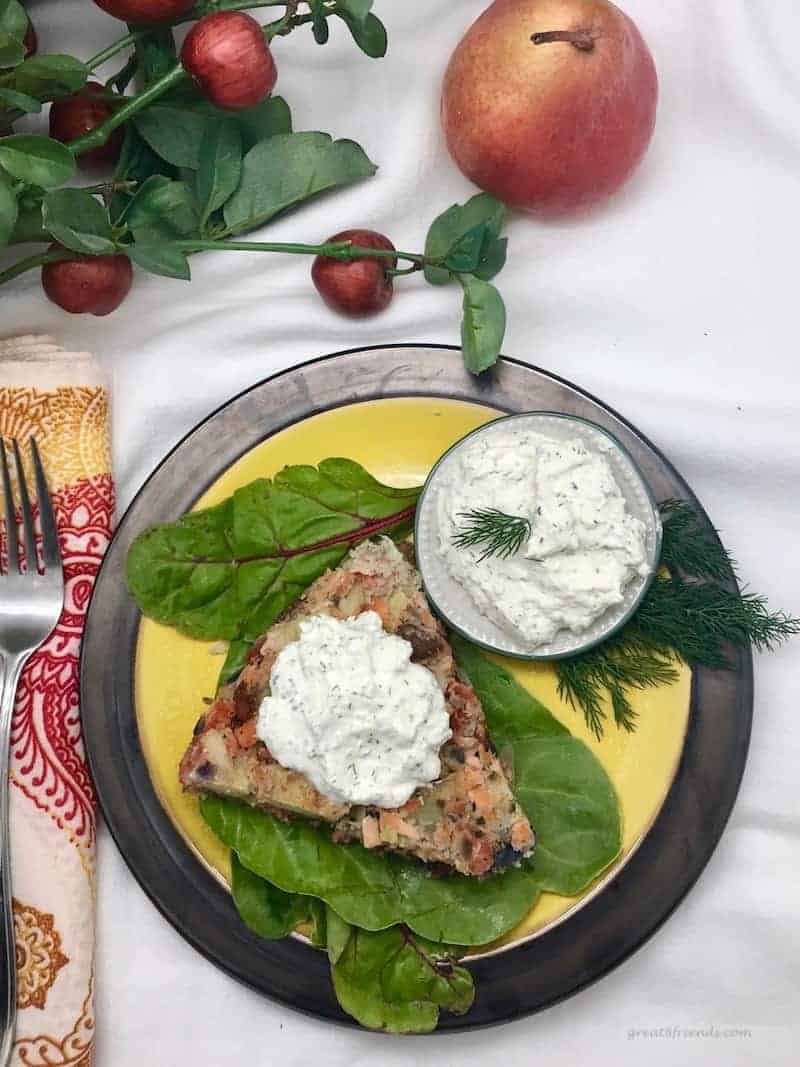 Overhead shot of black charger with a yellow plate containing some chard leaves topped with a wedge of salmon hash with a dollop of dill sauce. There is a small bowl of dill sauce to the side.