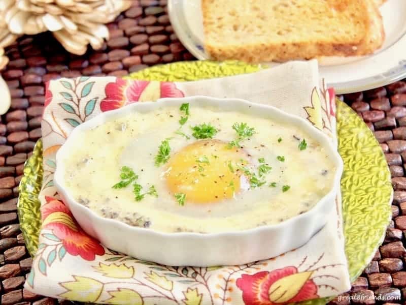 An Egg with cream sauce backed in a small oval ramekin sitting on a flowered napkin on a green glass plate.