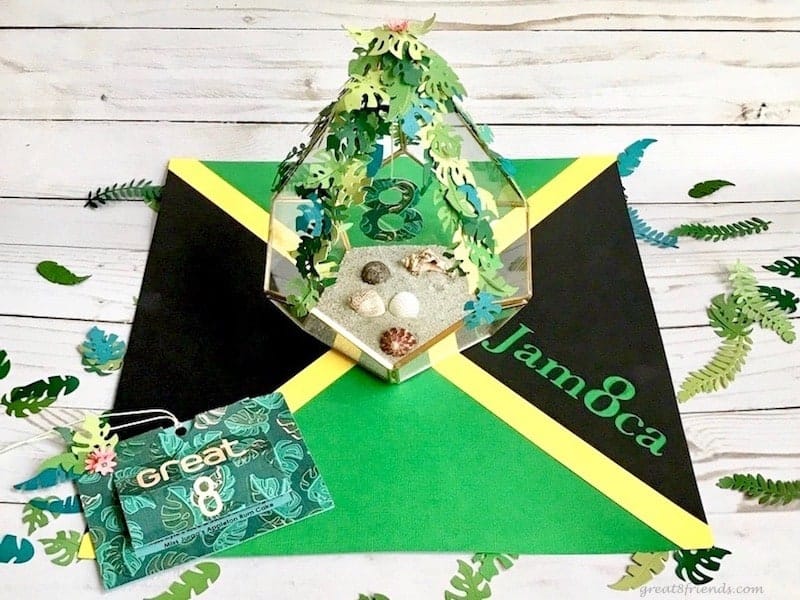 Jamaica theme dinner party invitaiton with the flag as a backdrop and a small terrarium with sand and shells and paper leaves.