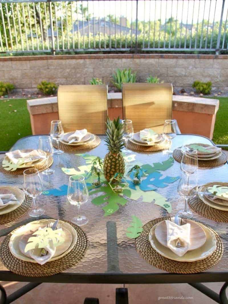 Outdoor table set with woven placemats, pineapple centerpiece sitting on cut out palm leaves.