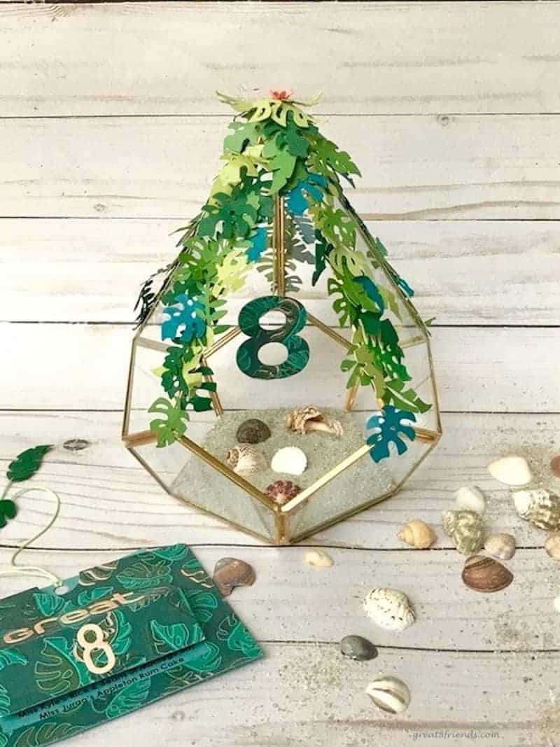 Close up of glass terrarium with sand and shells and paper leaves and a cut-out 8 hanging in the middle.