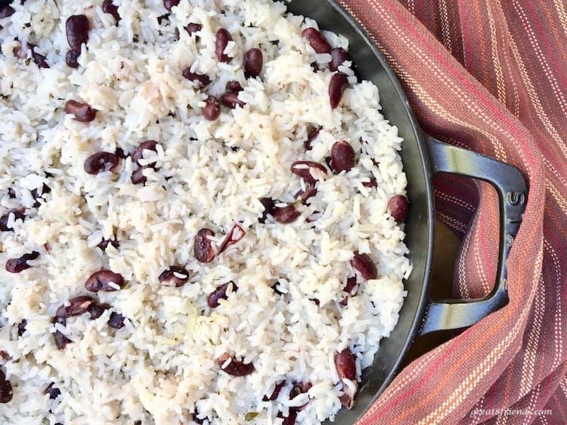 Jamaican rice and beans in a black skillet with a brown striped kitchen towel on the side.