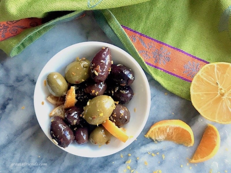 Overhead shot of a small white bowl of citrus marinated olives with a green towel and sliced lemon.
