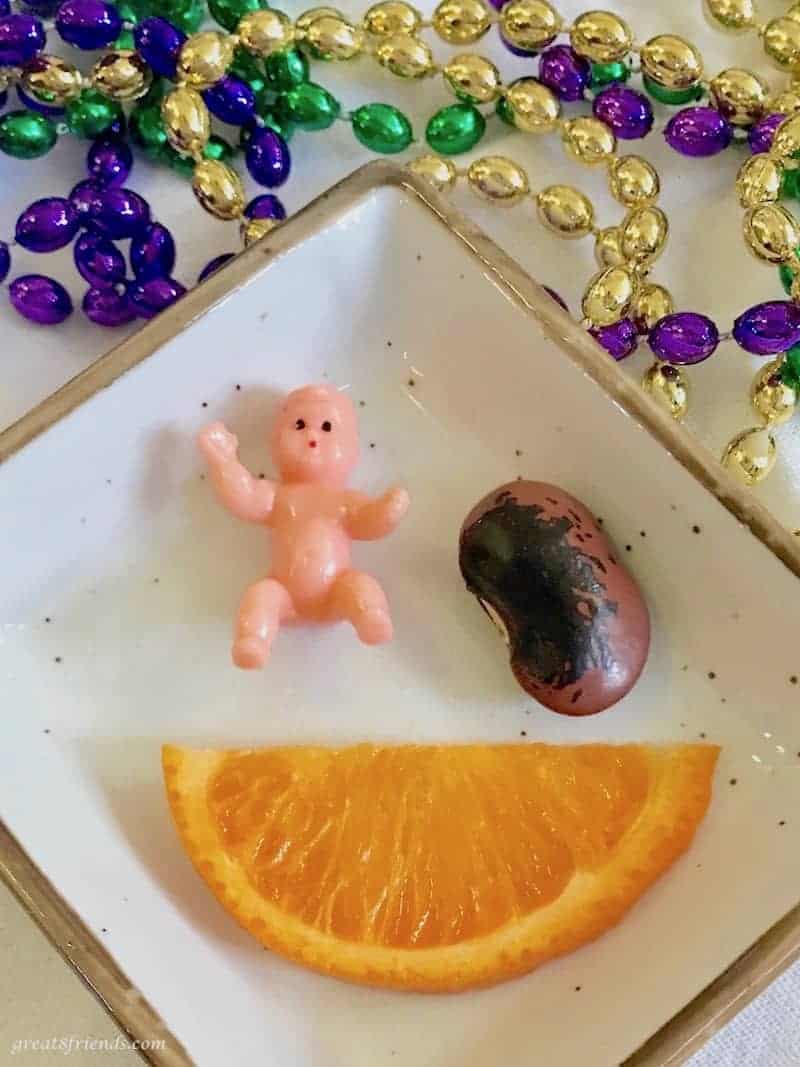 Plastic mini Baby, a bean and an orange slice on a square plate surrounded by Mardi Gras beads.