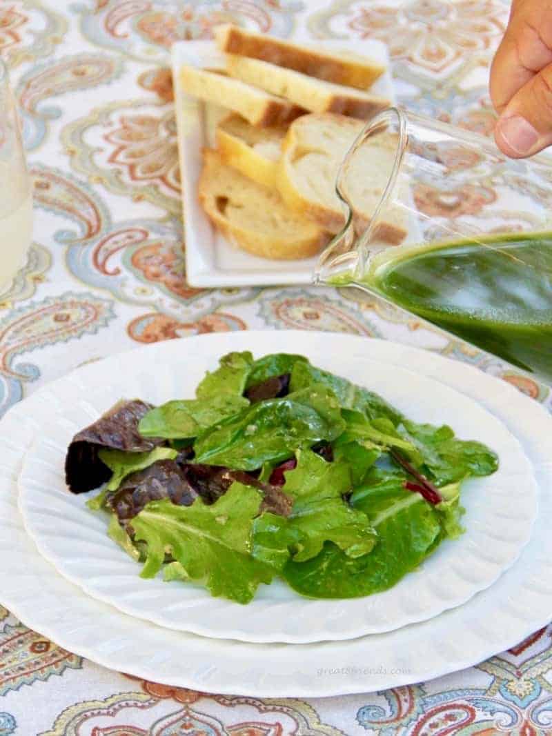 Green salad on a white plate with green arugula lemon salad dressing being poured on with sliced bread in the background.