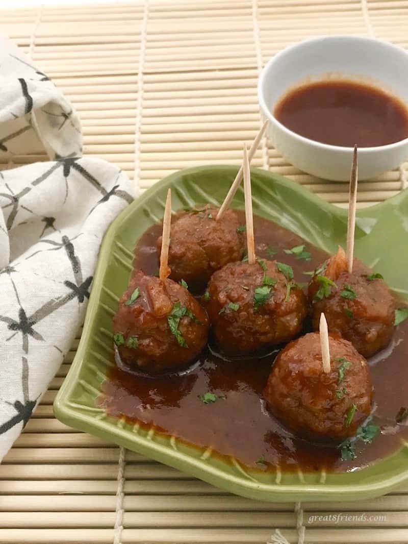 Five Hawaiian Meatballs on a green dish with toothpicks in each for eating and a bowl of extra sauce on the side.