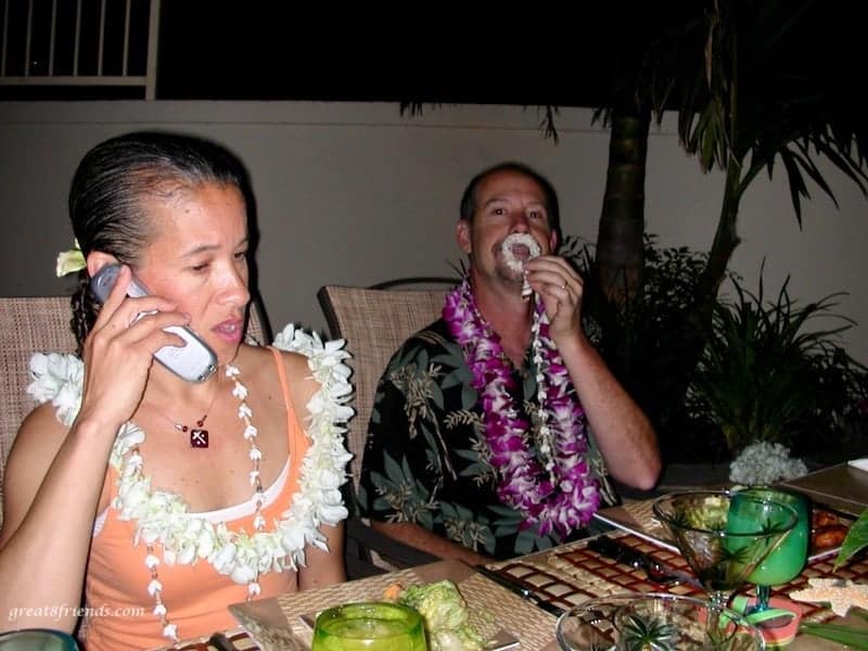 Two people (one on the phone) with Hawaiian leis at a backyard table set for dinner.