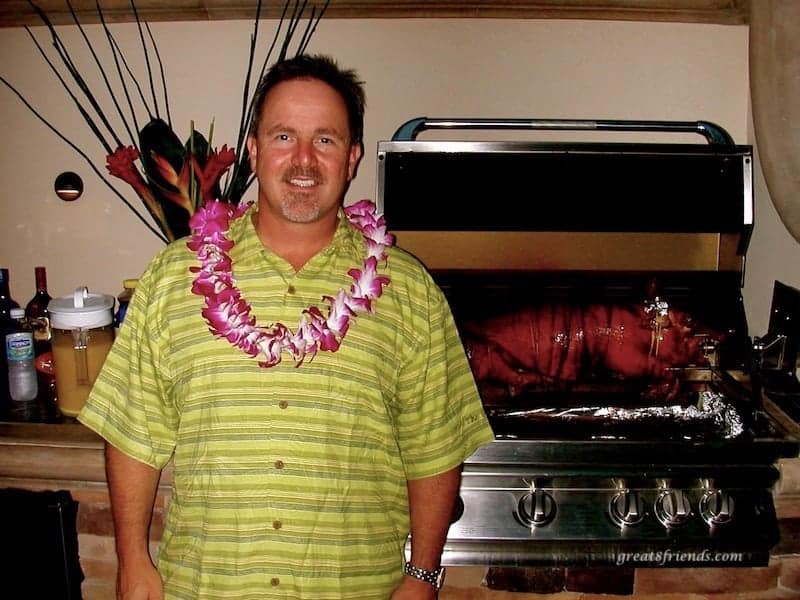 A man posing with a whole pig in the background roasting on a spit on a backyard grill.