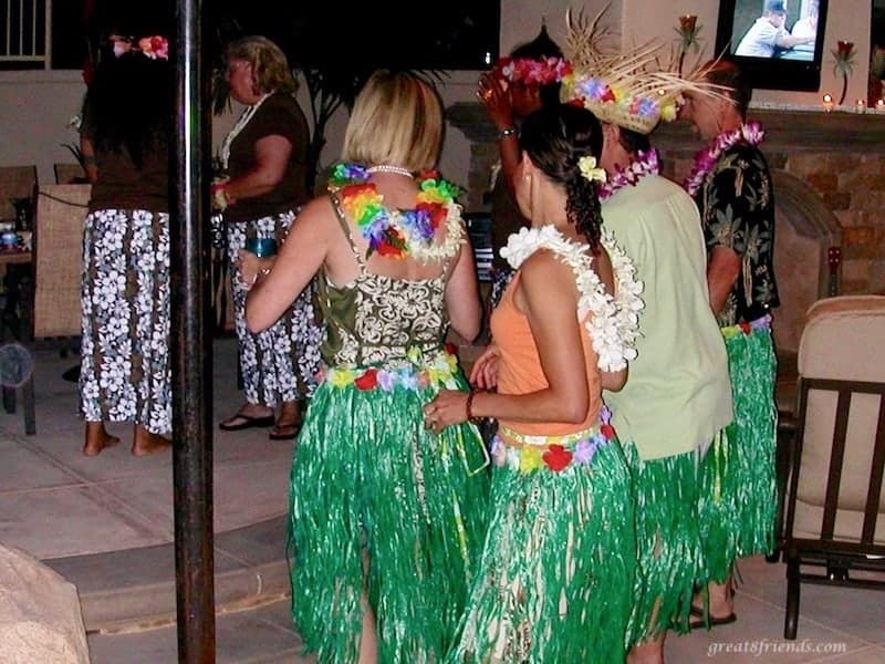 Two women and two men in green grass hula skirts learning the hula dance from two female instructors in flowered skirts.