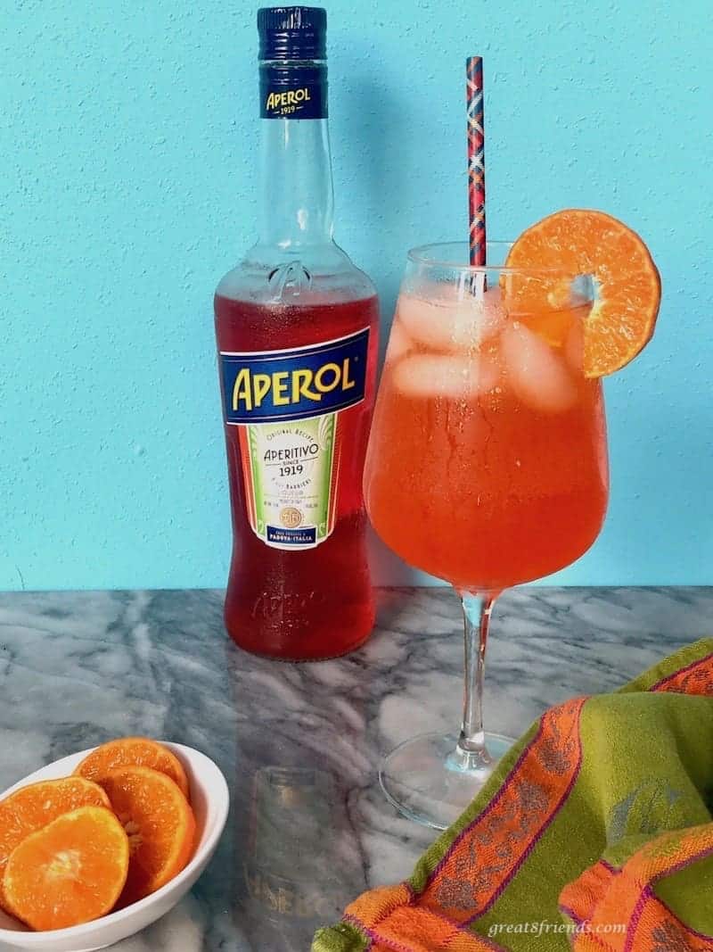 Aperol Spritz cocktail on a counter with a bottle of Aperol and sliced oranges in a bowl.