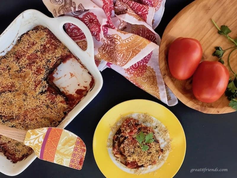 Pan of eggplant parmesan with one slice cut out on a small plate and whole tomatoes in a wooden bowl.