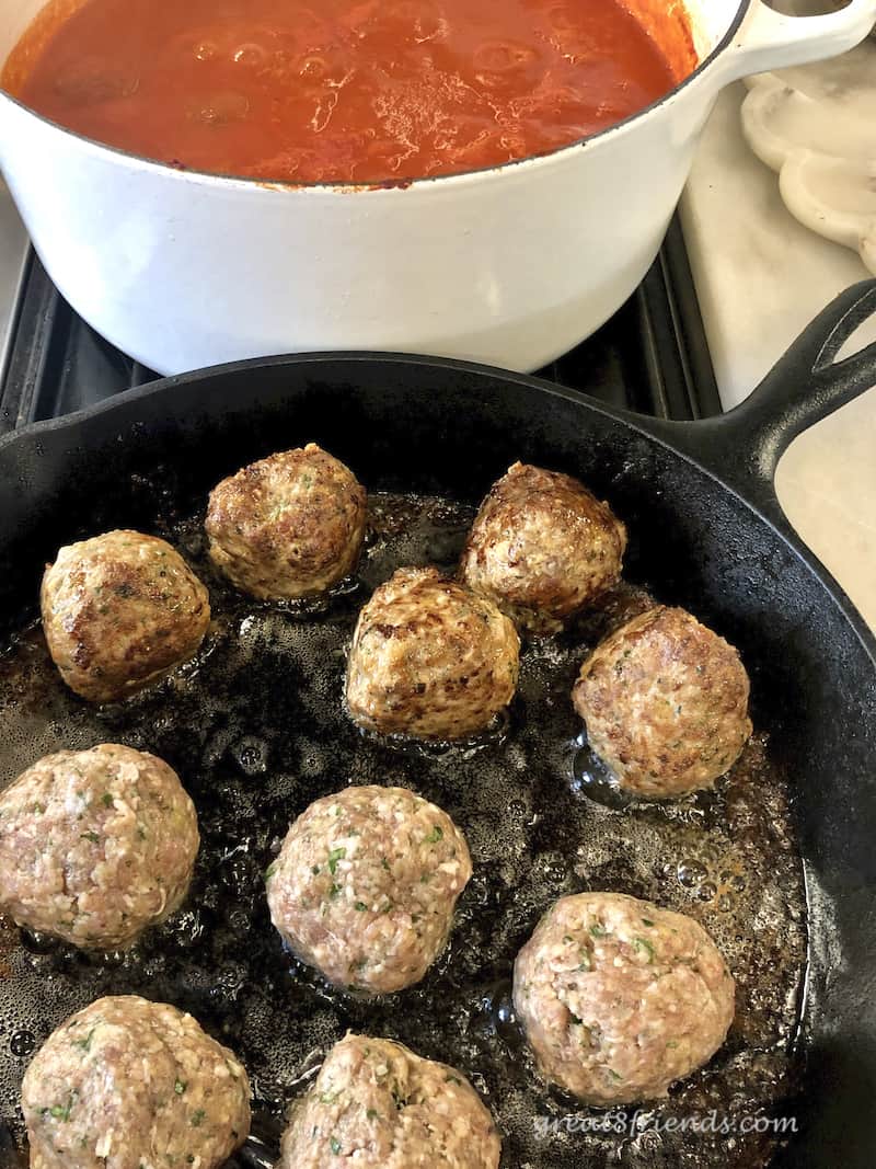 Meatballs frying in cast iron skillet with white pan of red sauce in the background.