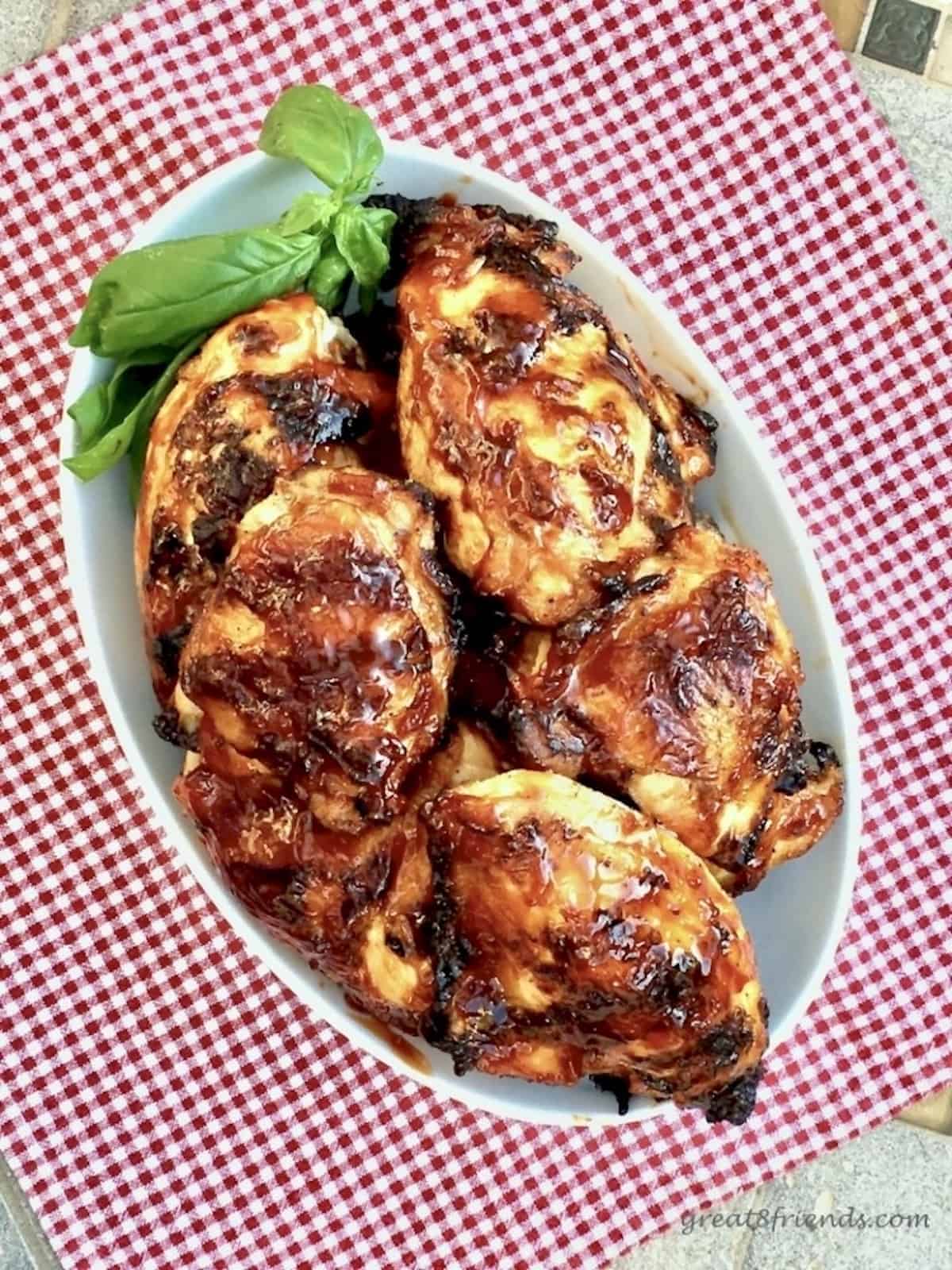 Barbecued Chicken pieces in an oval white dish sitting on a red and white gingham cloth with a basil leaf as garnish.
