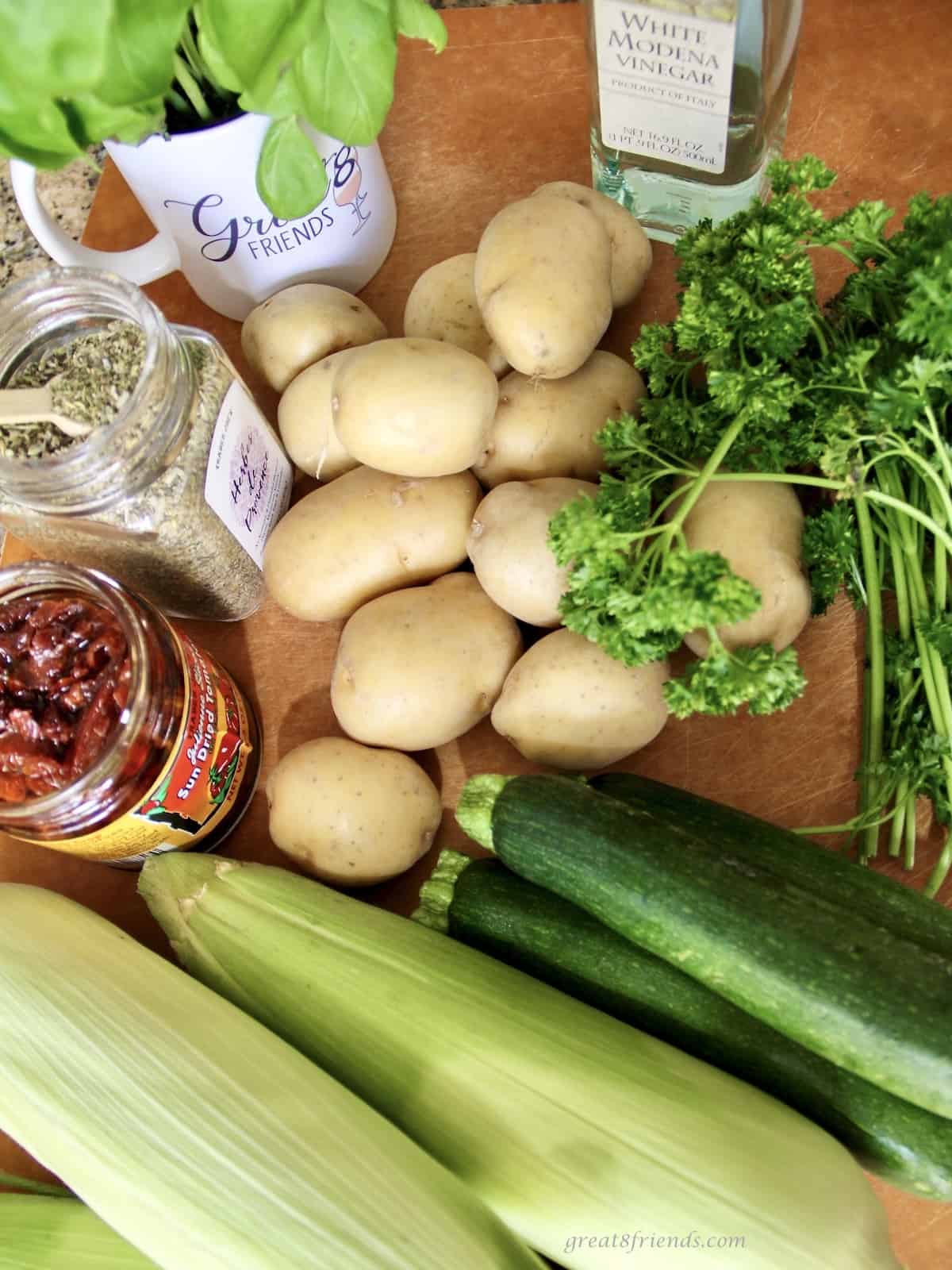 Overhead view of the ingredients in a salad including potatoes, zucchini, corn in the husk, basil, parsley, herbs de Provence, sun-dried tomatoes and vinegar.