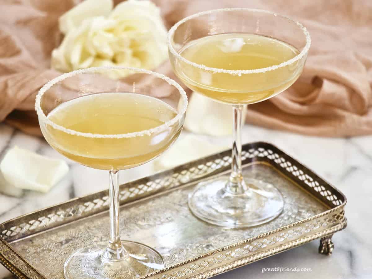 Two sidecar cocktails in coupe glasses on a silver tray with a white rose on pink in the background.