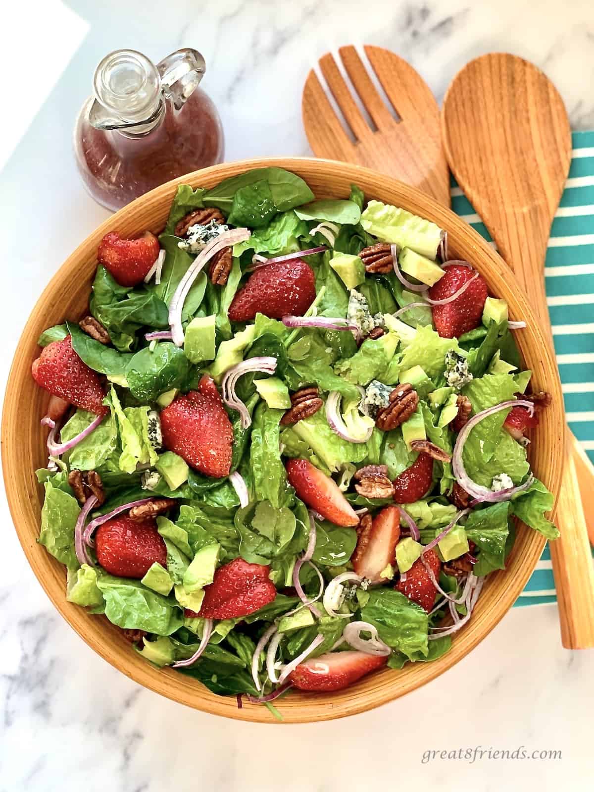 Overhead shot of a salad of vegetables, blue cheese, candied pecans in a wooden bowl with salad servers and a blue and white striped napkin.