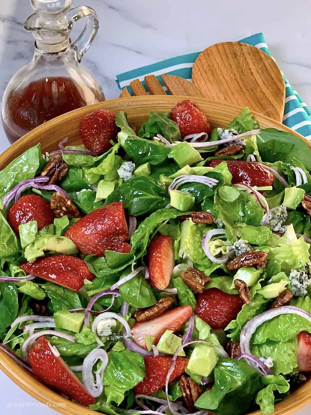 A mixed green salad with strawberries, pecans, and red onion in a wooden bowl.