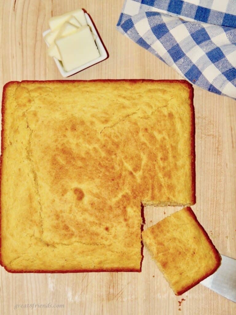 Overhead shot of a square cornbread with one piece cut with butter and a blue checked napkin on the side.