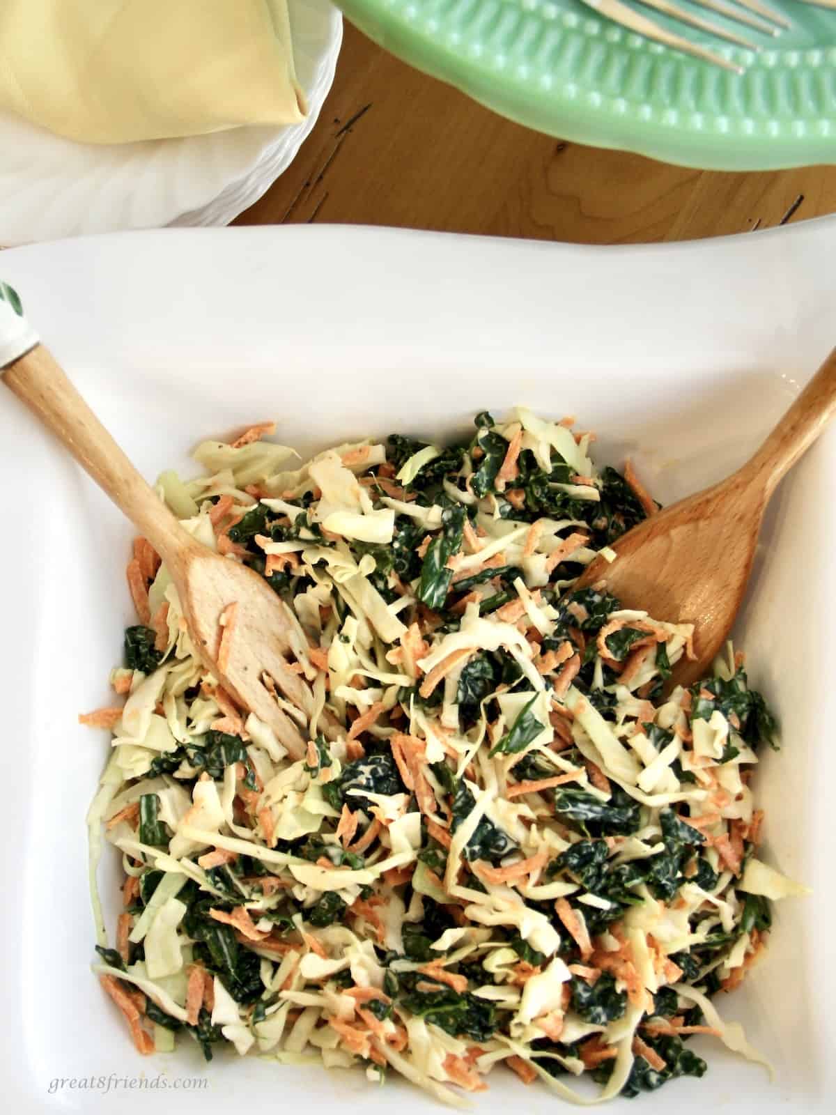 Kale slaw salad in a white square bowl with a wooden salad serving spoon and fork.