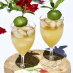 Two Mai Tais in stemmed iced tea glasses on a round wooden board with red flowers in the background.