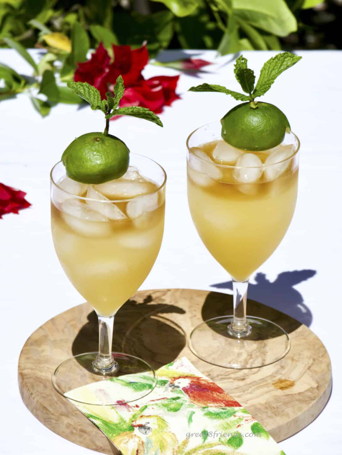 Two Mai Tais in stemmed iced tea glasses on a round wooden board with red flowers in the background.