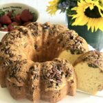 Rum Bundt cake on a cake stand with one piece sliced with whole strawberries and sunflowers in the background.