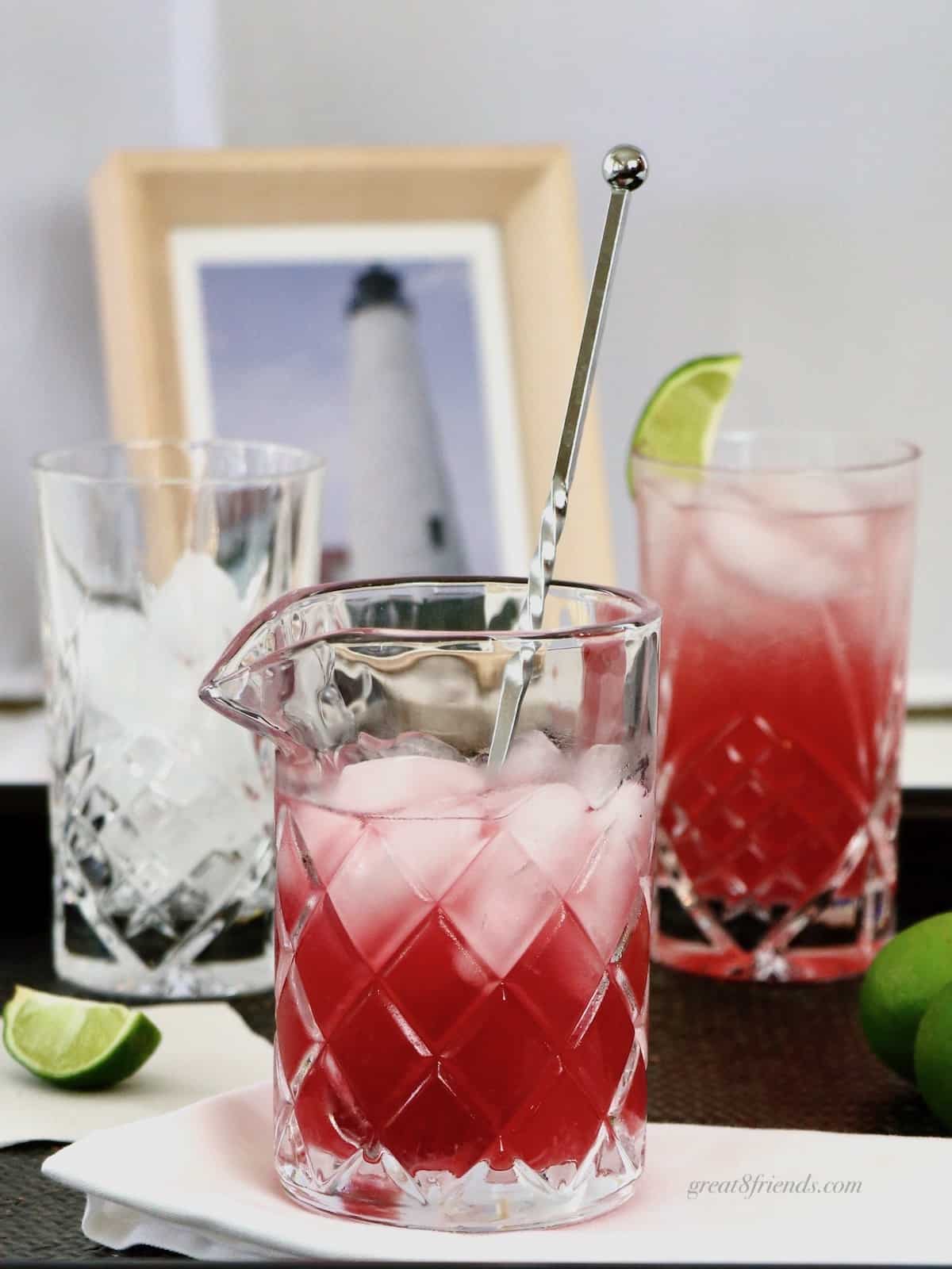 Clear glass pitcher with ice and Nantucket Breeze Cocktail with a spoon, and a highball glass with cocktail. A picture of a lighthouse in the background.