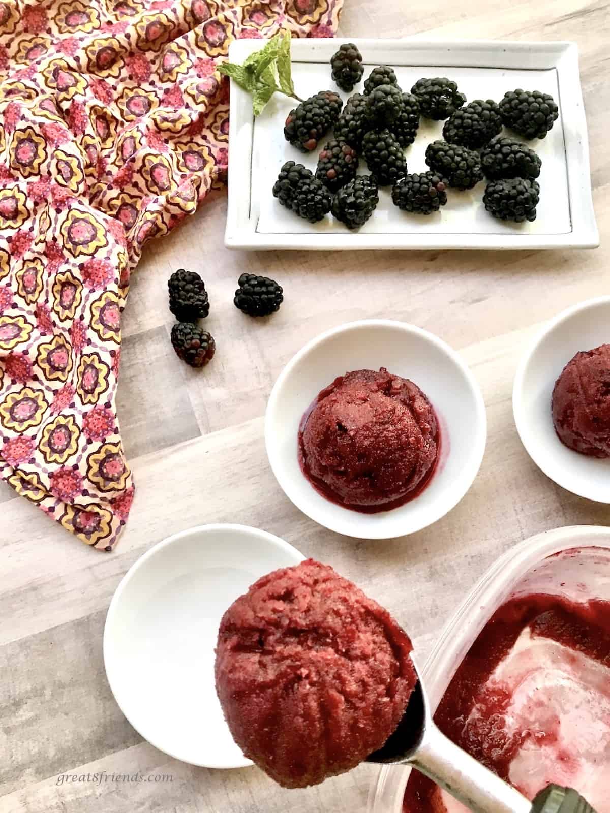 Italian ice scoops in white bowls one scoop in the ice cream scoop with blackberries on the side.