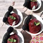 Four scoops of blackberry ice with fresh blackberries on a stick served on the side of the bowl.